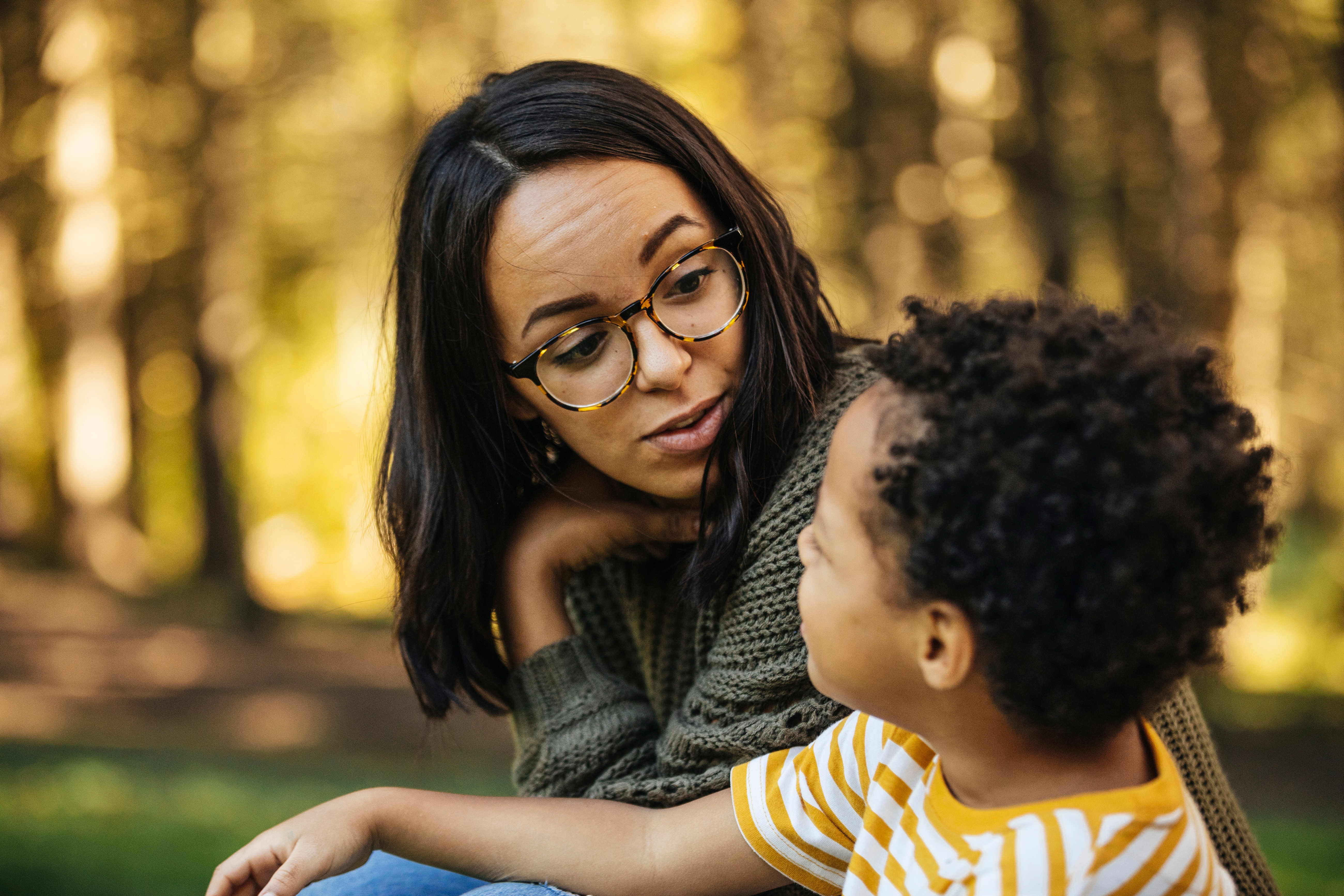 Woman in glasses and sweater crouches to converse with a young child in a striped shirt in a forest-like setting