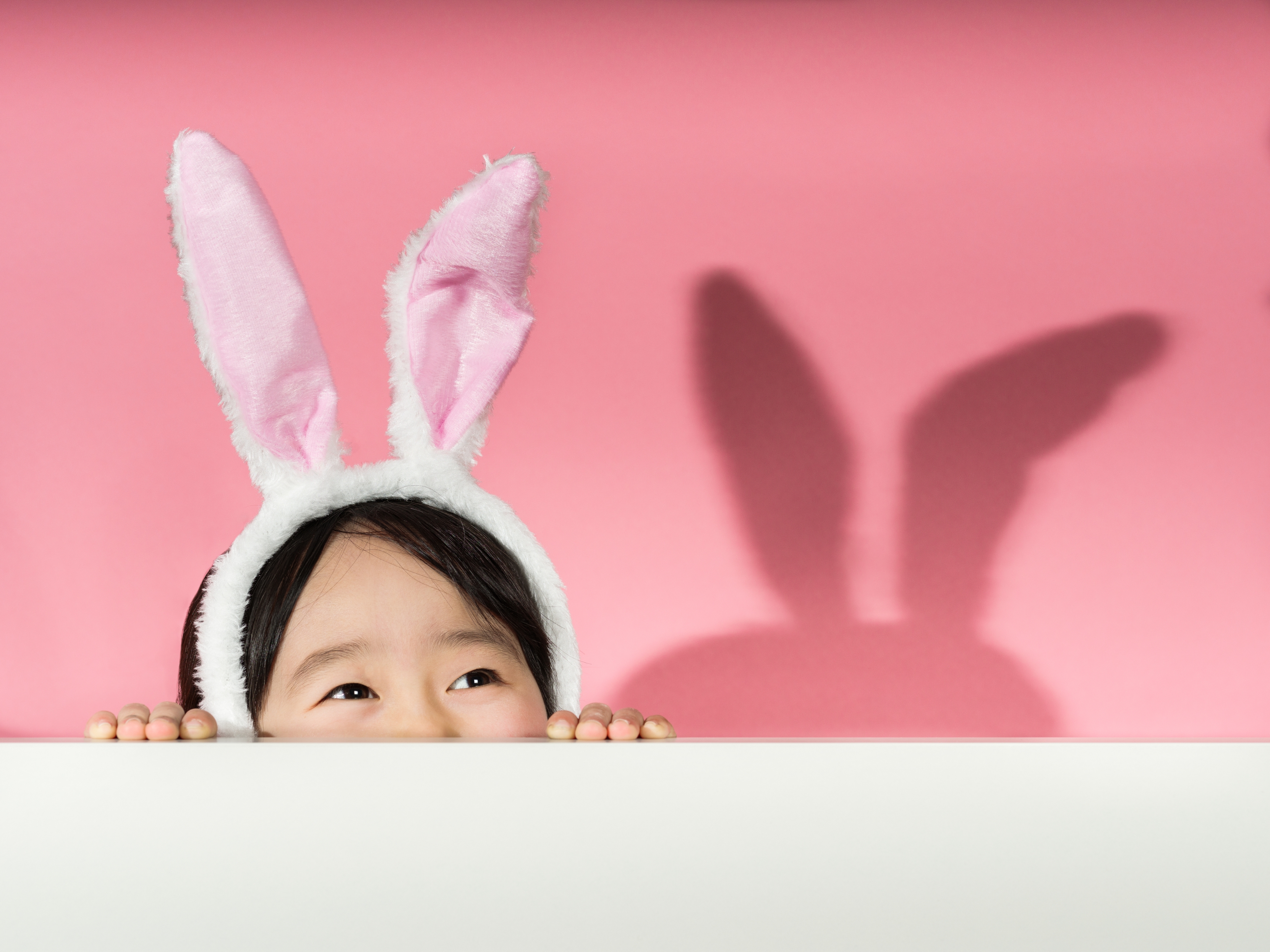 Child peeking over a surface wearing bunny ears headband, casting a bunny-shaped shadow on the wall