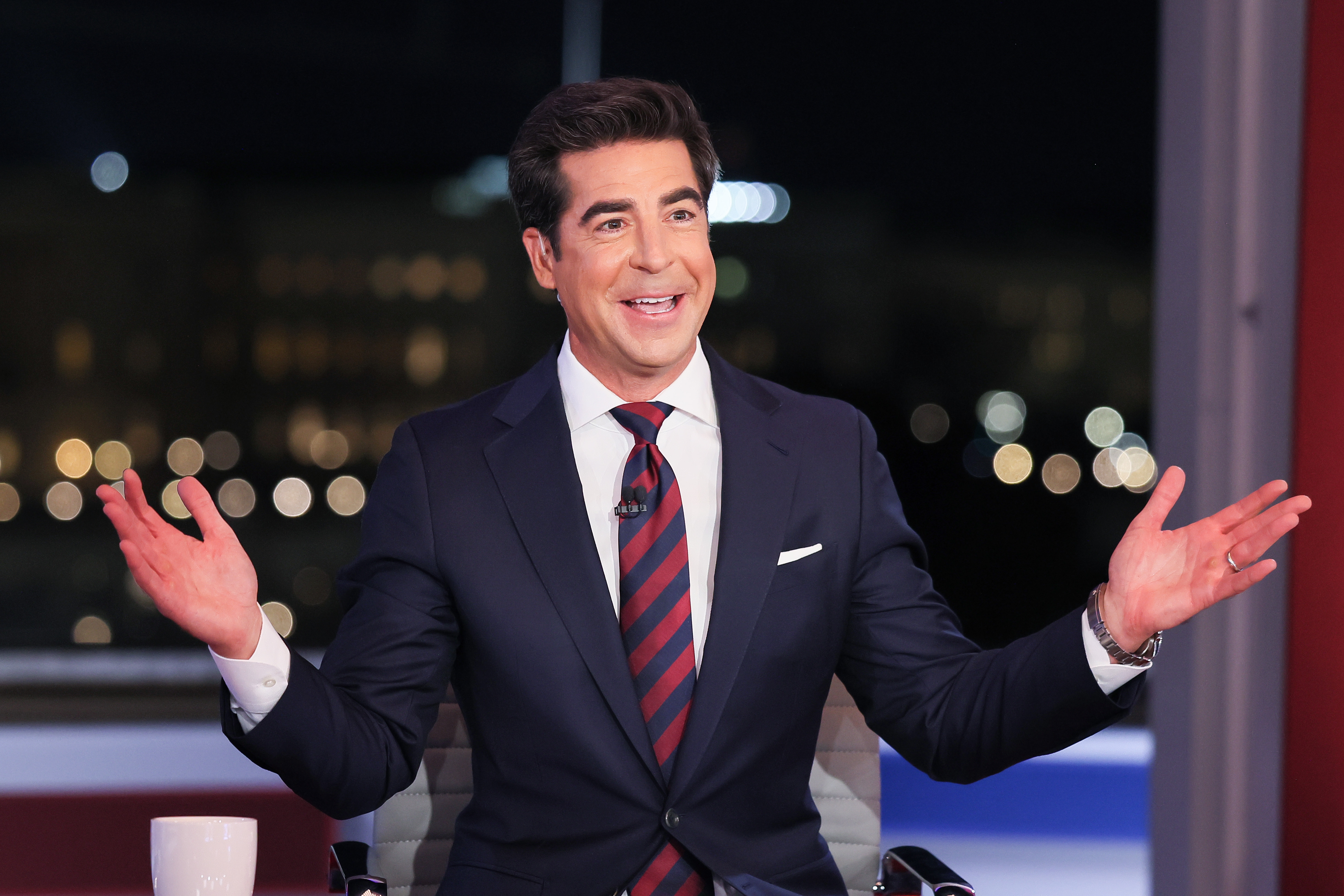Jesse Watters in a suit and striped tie gestures with open arms, sitting in a studio with blurred lights in the background