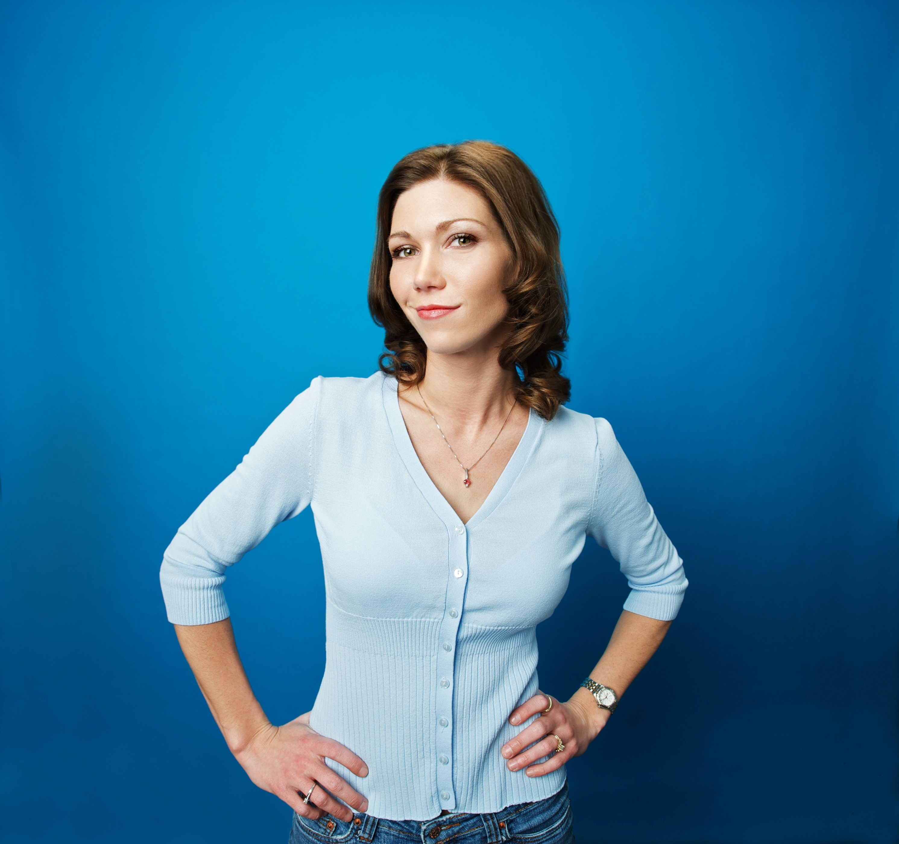 Person standing confidently with hands on hips, wearing a buttoned top and jeans against a plain background