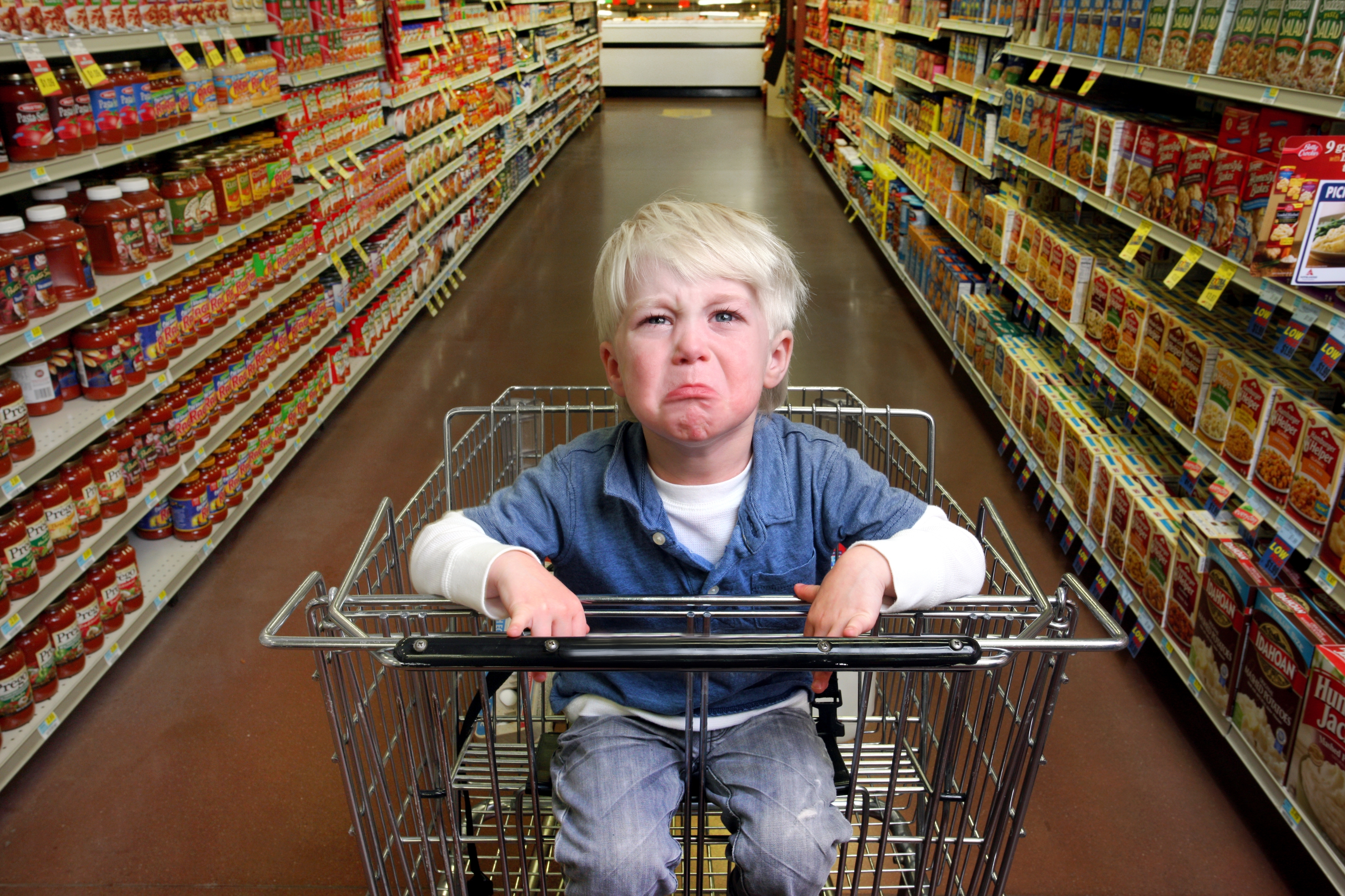 A crying child in a shopping cart inside a grocery aisle filled with various packaged products