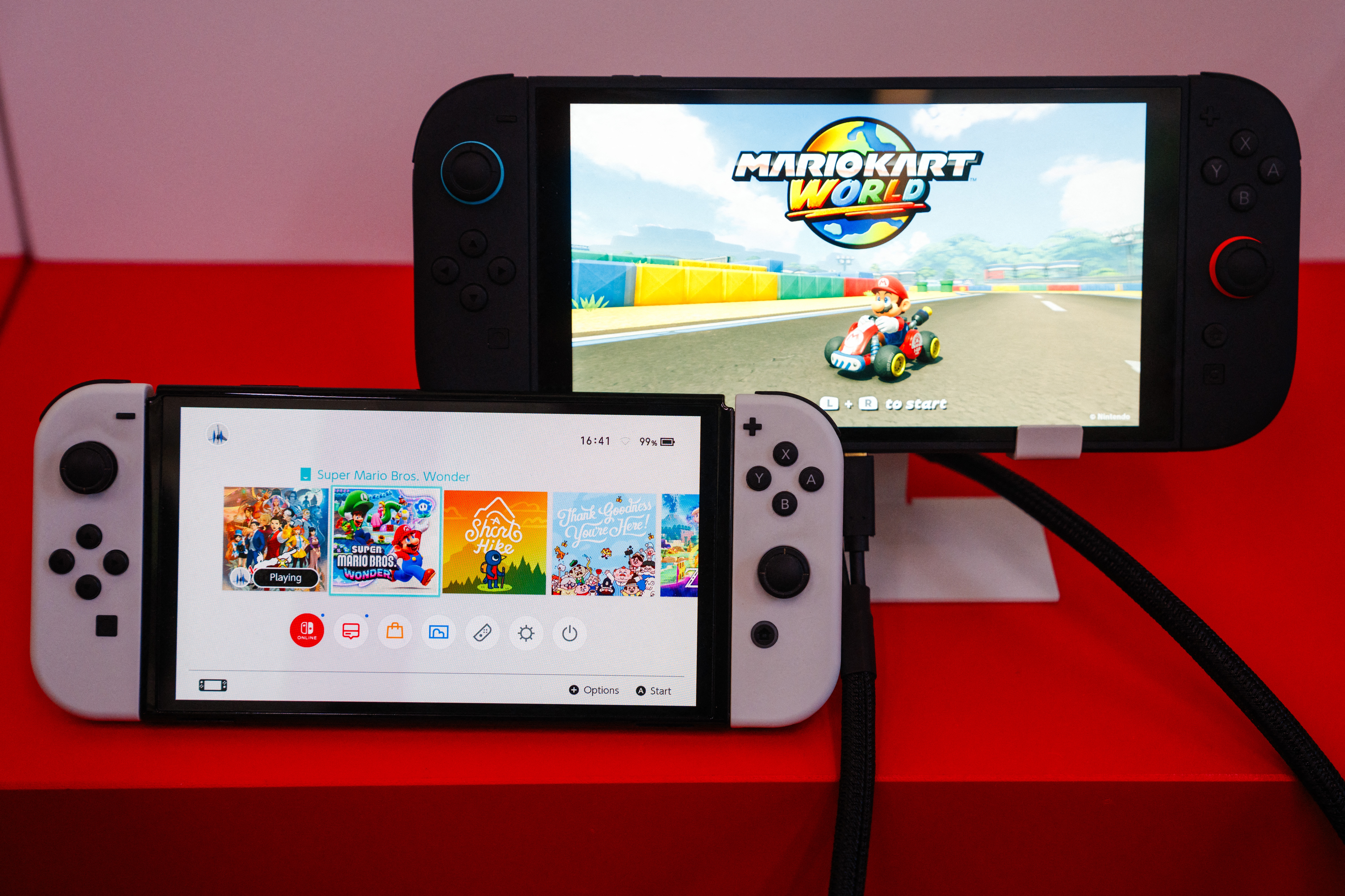 Two handheld gaming consoles displaying games; the front shows a selection menu, and the back displays "Mario Kart World" on-screen