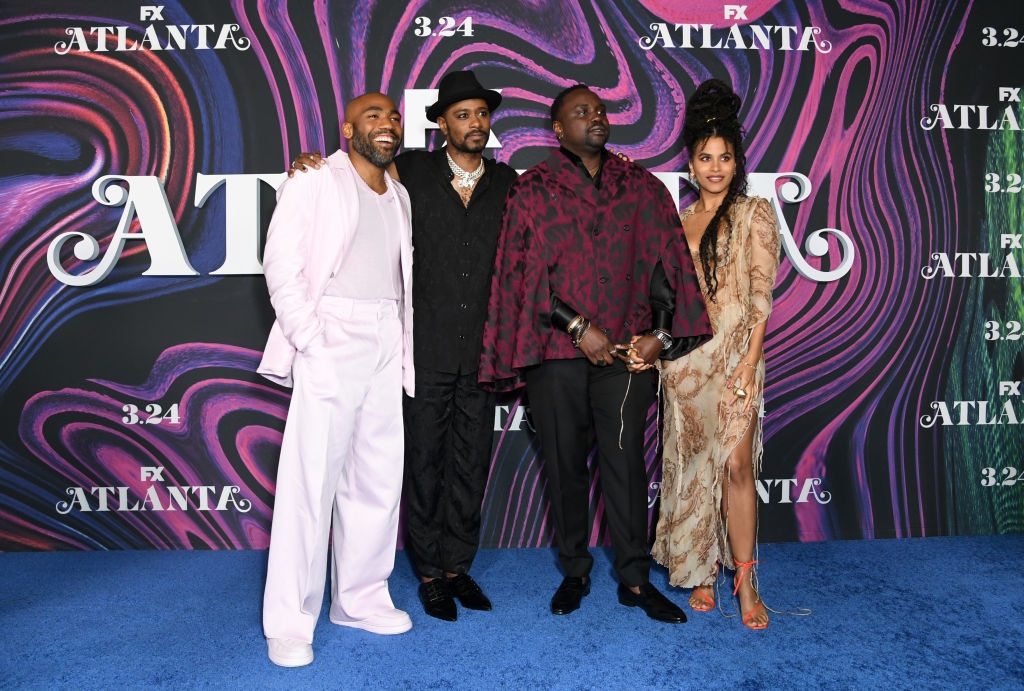 Four people pose on the blue carpet at an &quot;Atlanta&quot; event; one wears a pink suit, others in hats and stylish outfits, standing against a vibrant backdrop