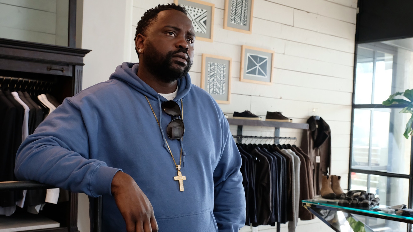 Person in a casual hoodie with a cross necklace, leaning against a stylish boutique interior with clothing racks in the background