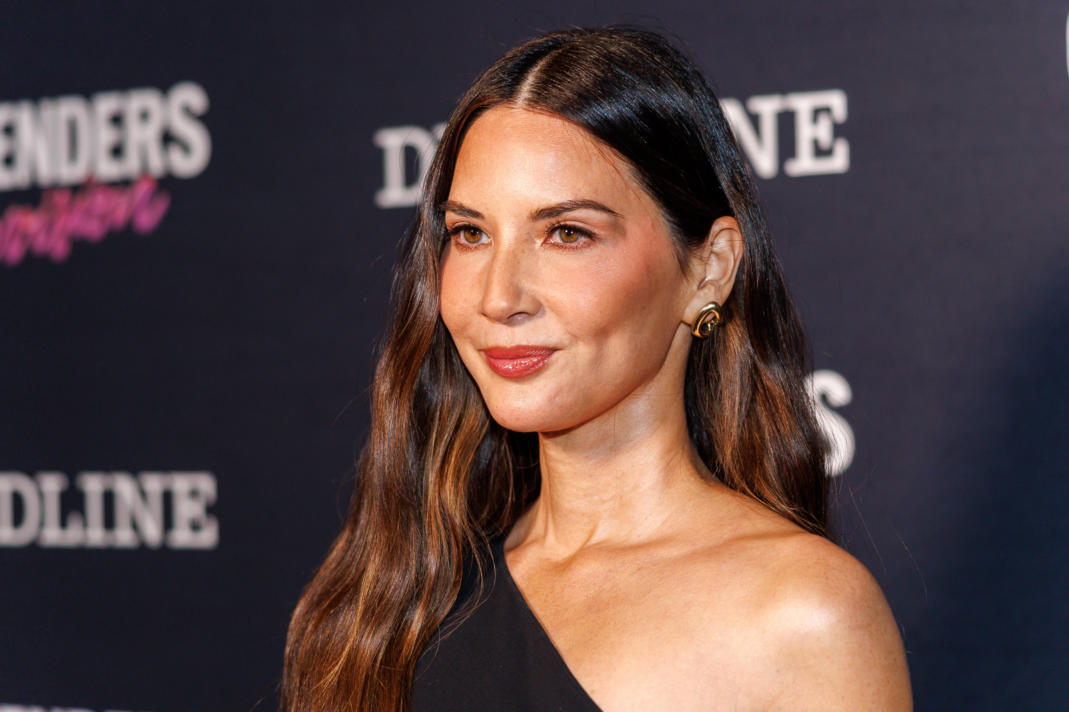 Olivia Munn in elegant, asymmetrical gown at a media event, smiling for photos on the carpet