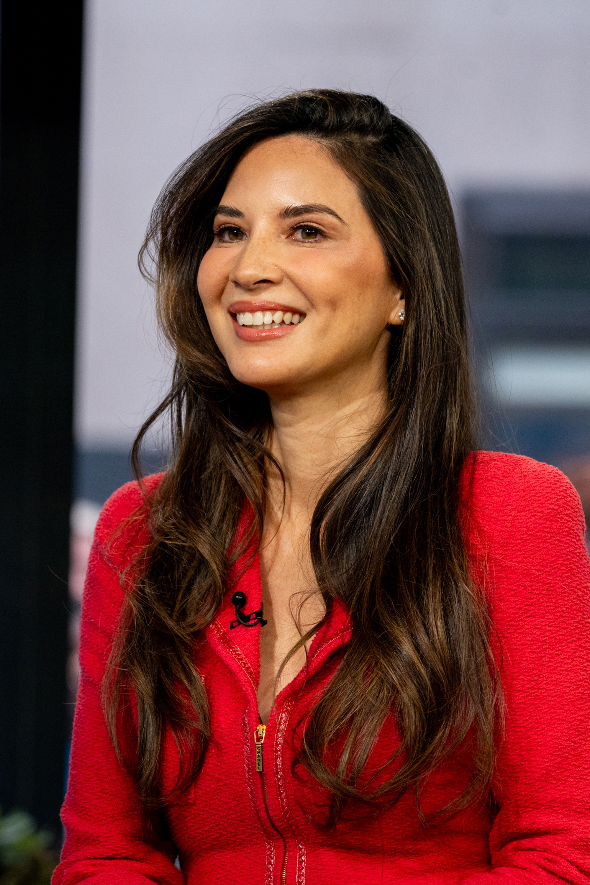 Olivia Munn smiles while wearing a fitted, textured outfit with a zipper detail during a public appearance