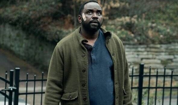 Man outdoors, with a thoughtful expression, wearing a green jacket over a blue shirt, and a bandage on his cheek