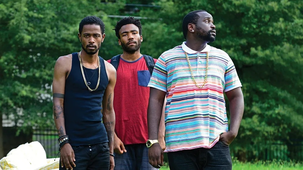 Three men stand outside; one wears a tank top, another a red T-shirt, and the last a striped shirt. They are from a TV show