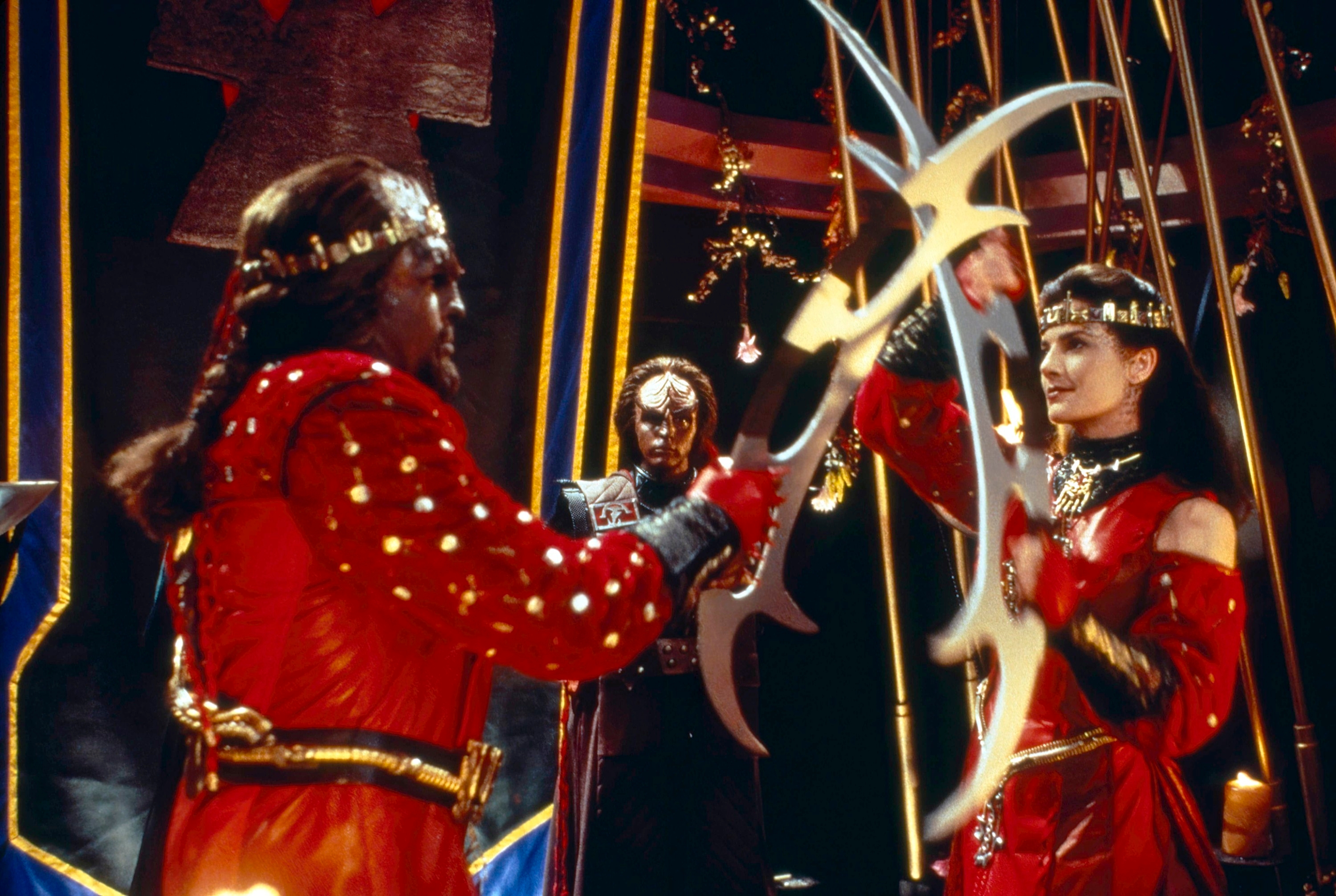 Sci-fi characters in ornate, red costumes duel with futuristic weapons on a set resembling a spaceship interior