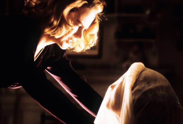 A person with short hair leans over a veiled figure in an atmospheric scene, suggesting mystery or tension, related to film or TV