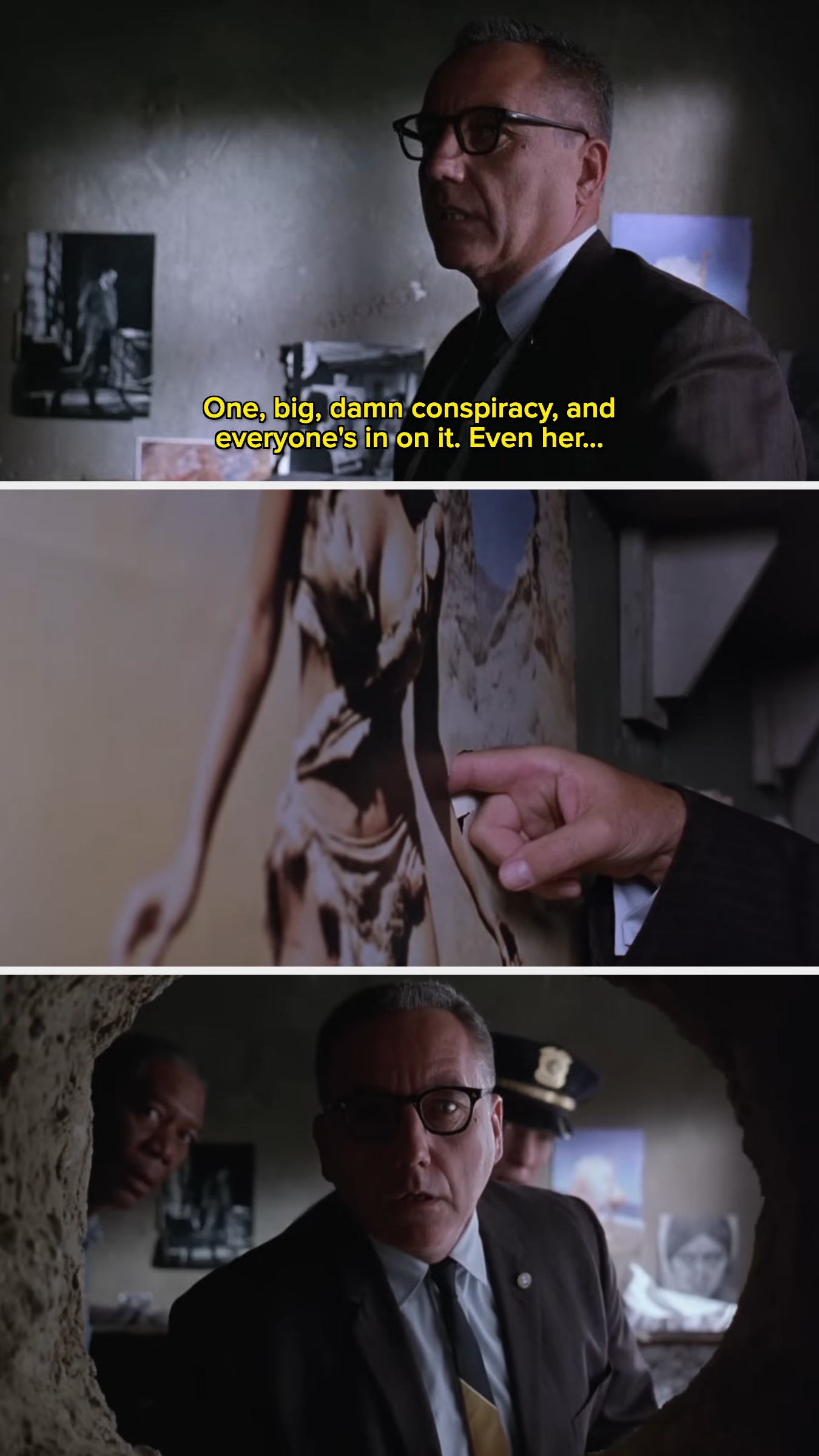 A man in a suit and glasses discusses a conspiracy, pointing at a photo on a wall, with another man partially in view