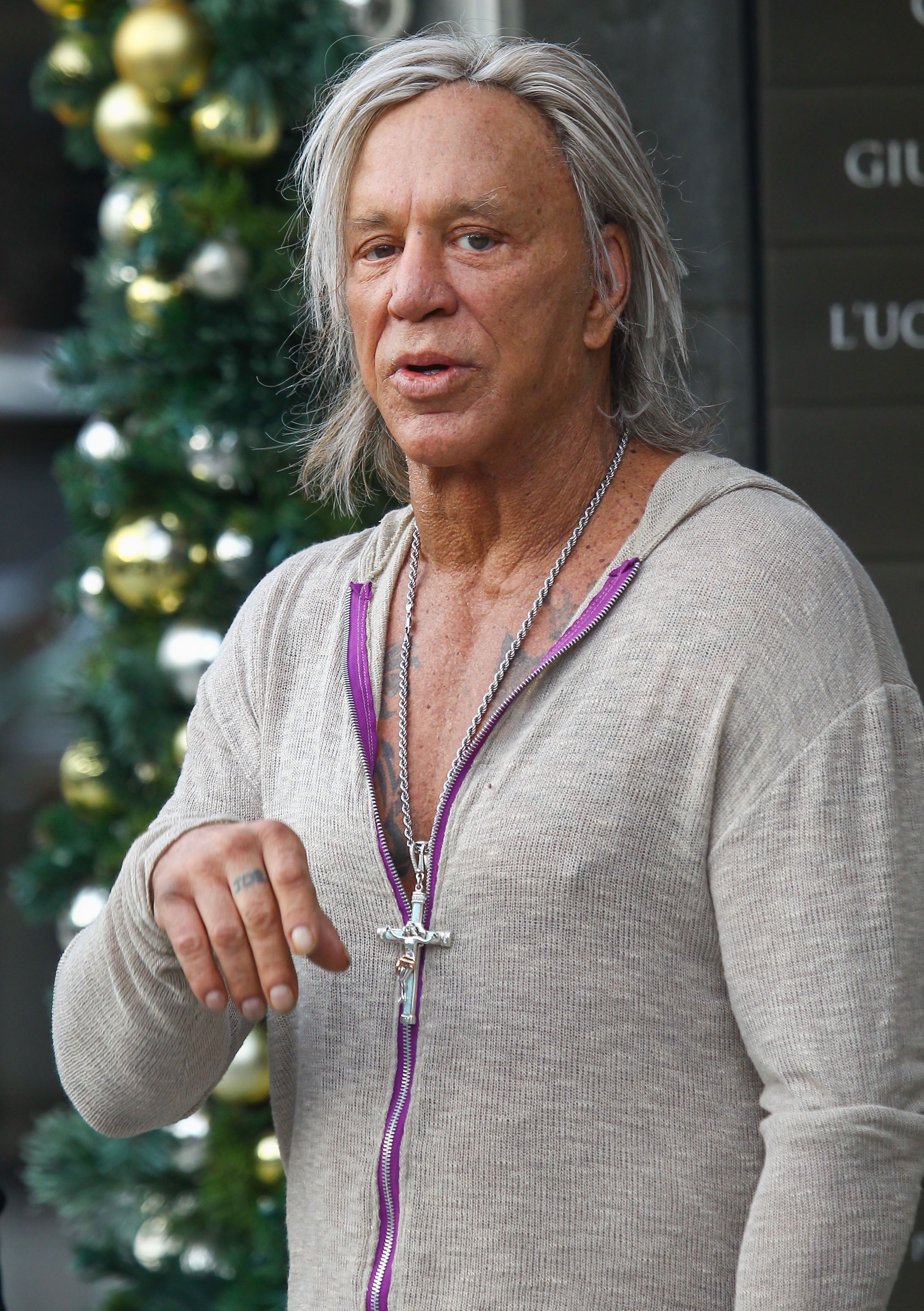 Mickey Rourke wearing a casual hoodie and a cross necklace, standing outside with festive decorations in the background
