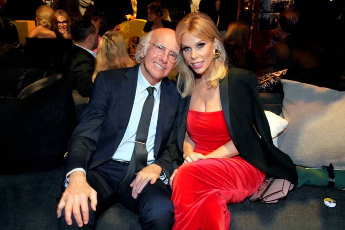 Larry David in a suit and Cheryl Hines in a strapless gown and blazer sit together on a couch at an event, smiling at the camera