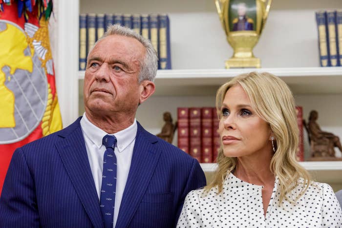 Robert F. Kennedy Jr. and Cheryl Hines stand together in a formal setting, both in business attire. Books and a decorative flag are visible in the background