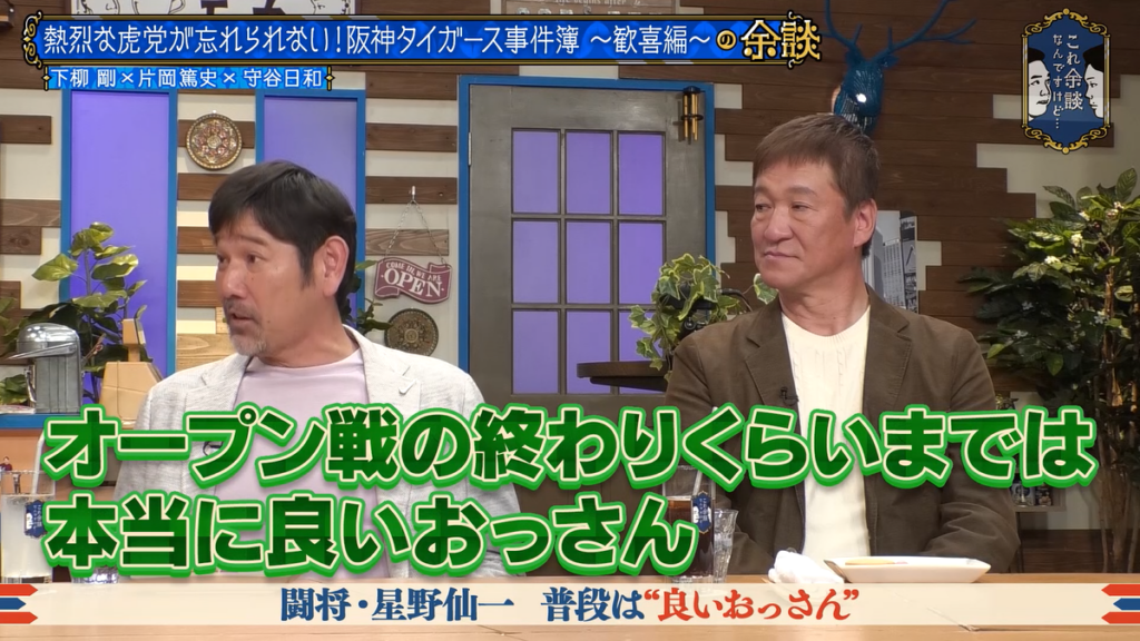 画像には、トーク番組に出演中の2人の男性が写っています。セットは居間風のインテリアです。