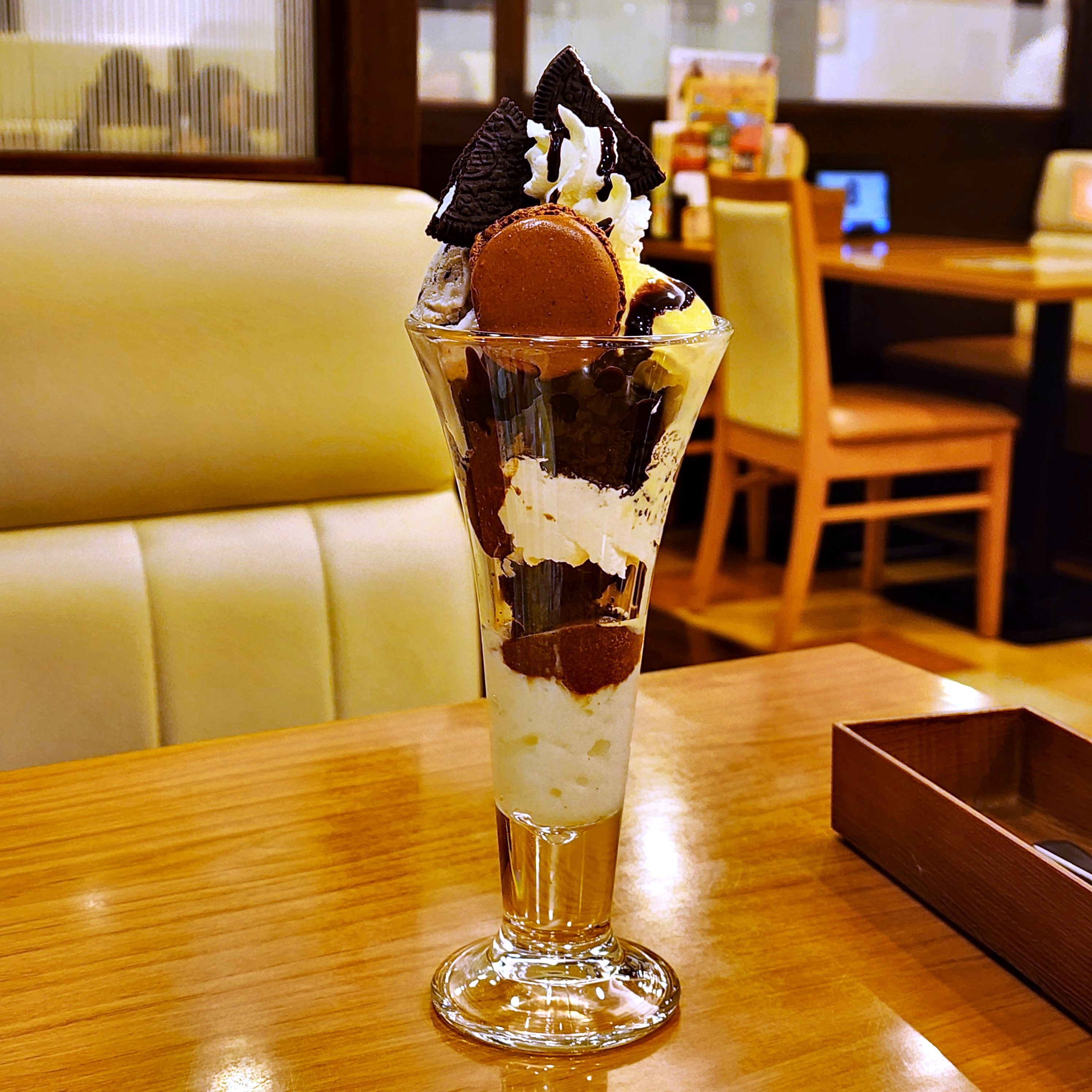 高いグラスに盛られたパフェ、クッキーやホイップクリーム、チョコレートがトッピングされています。背景はカフェの内装です。