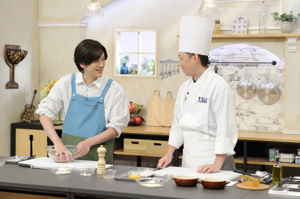 男性二人がキッチンで料理中。左はエプロン姿で生地を捏ね、右はシェフ帽をかぶった料理人。