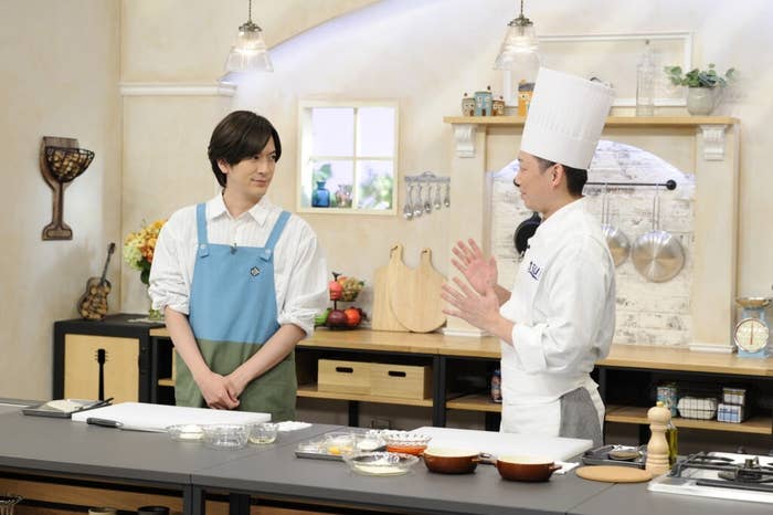 キッチンにいるシェフ服姿のシェフと、青いエプロンを着た男性が会話している。