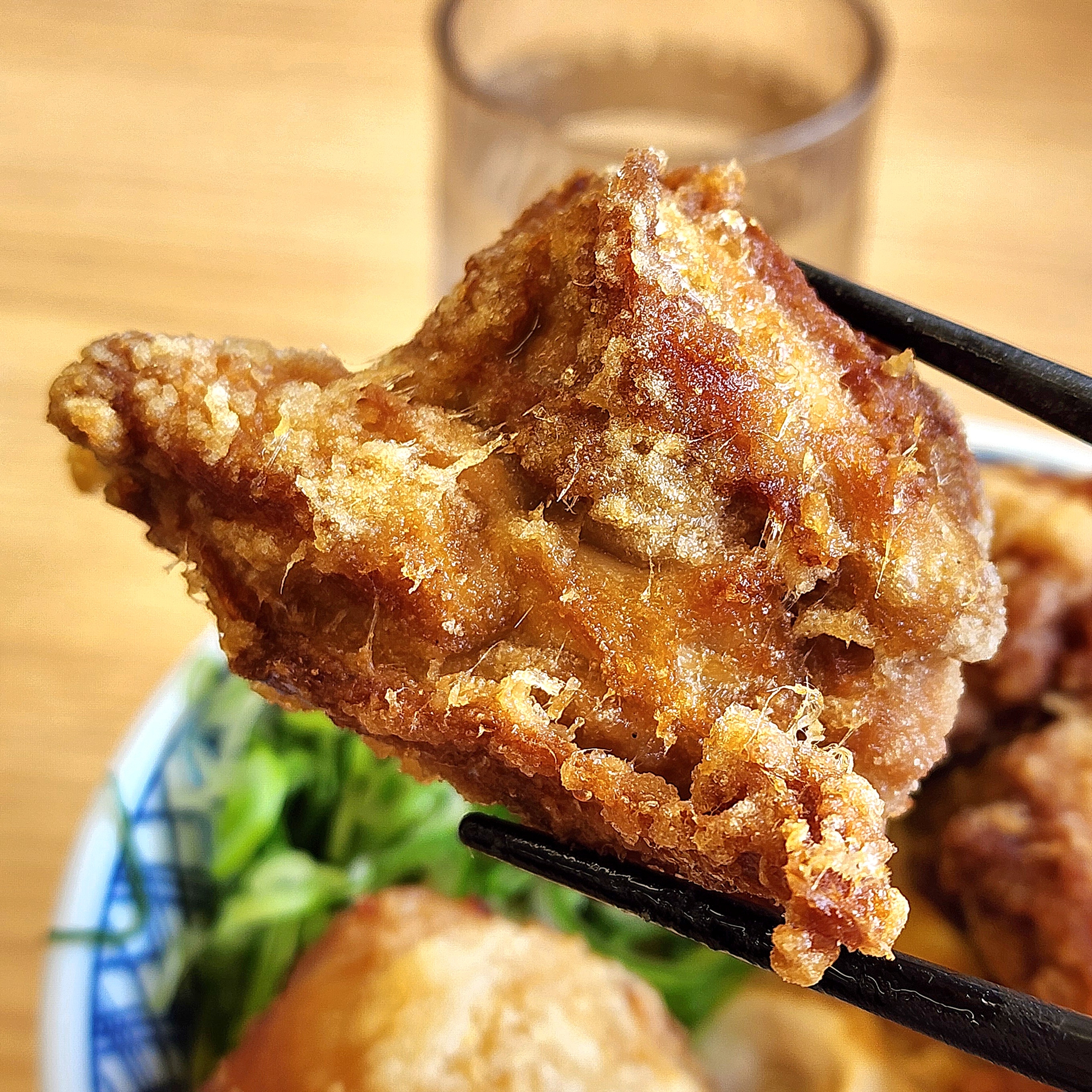 チキンのから揚げが箸で持ち上げられている。後ろにサラダと飲み物が見える。