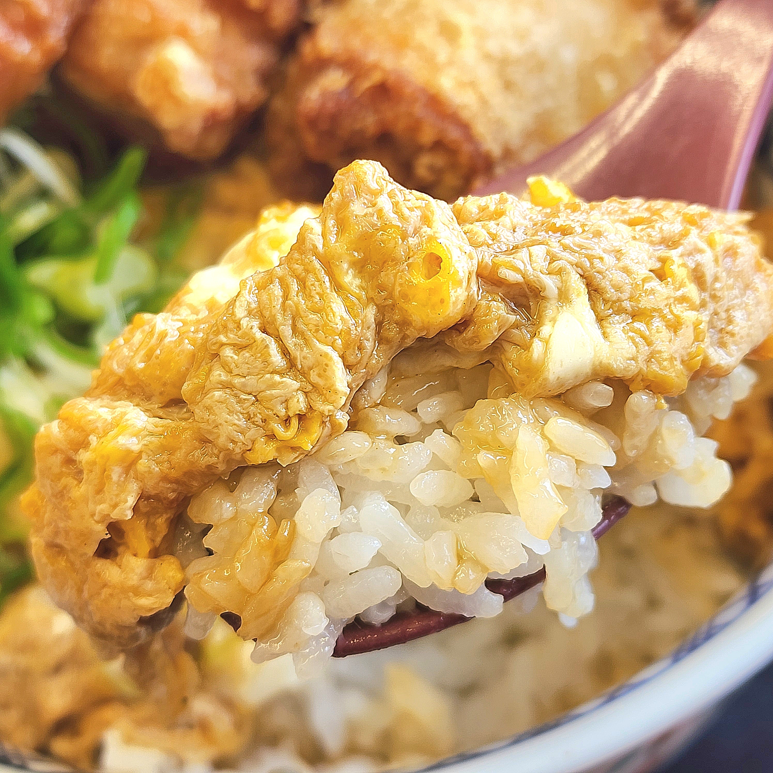 スプーンですくった卵とじご飯のクローズアップ。後ろに野菜や揚げ物も見える。