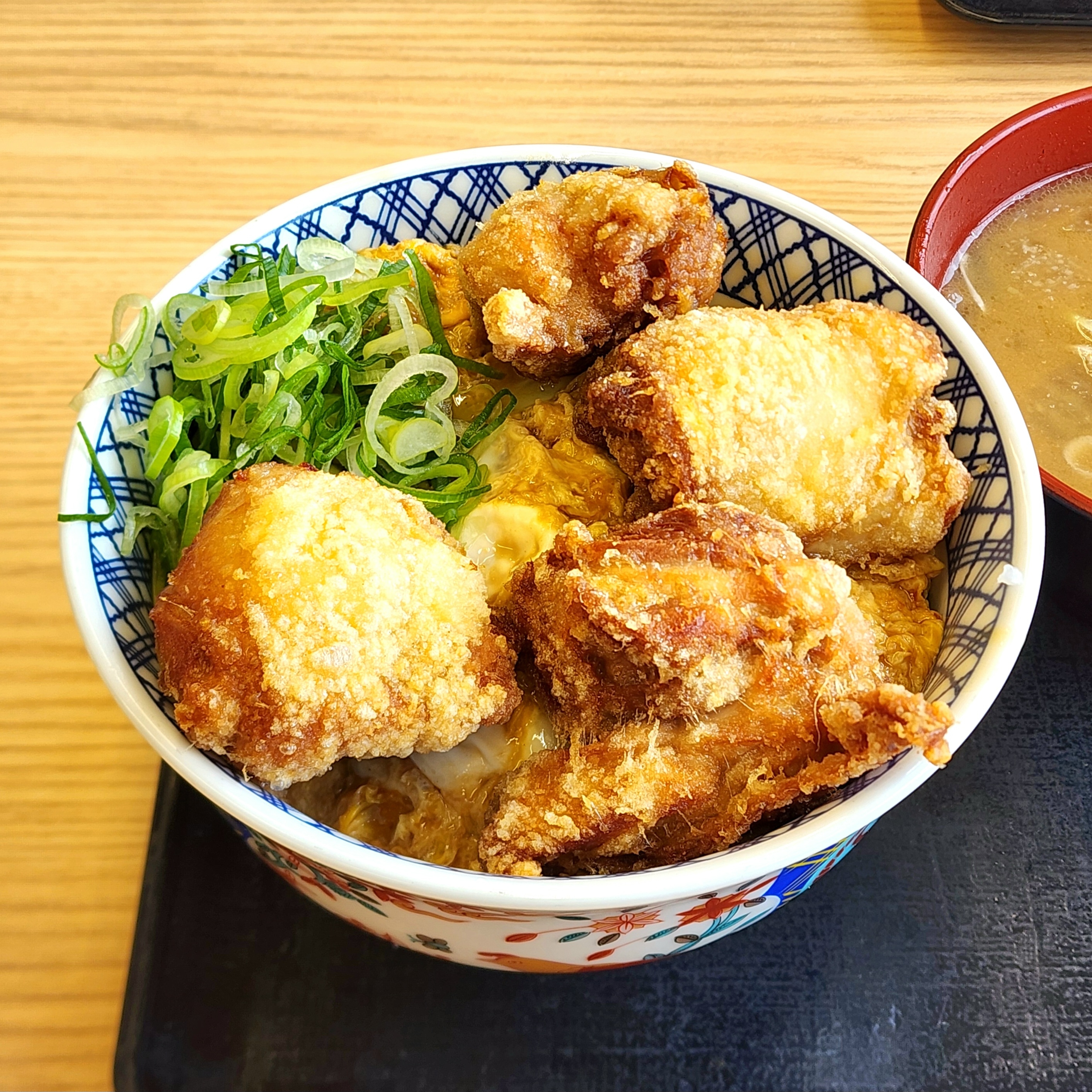 唐揚げが乗った美味しそうな丼ぶり、ネギがトッピングされている。隣に味噌汁の小鉢もある。