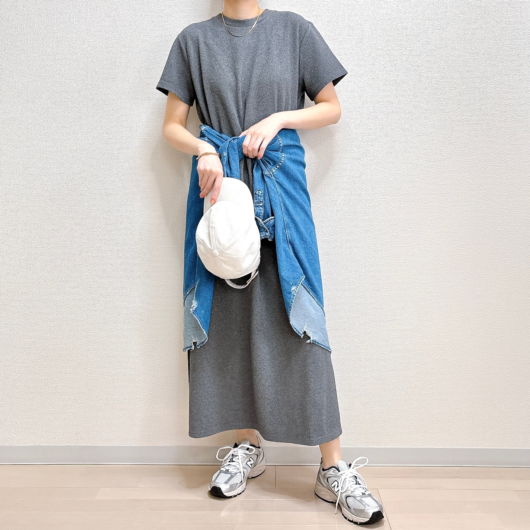 女性がシンプルなグレーのワンピースにデニムジャケットを腰に巻き、スニーカーを履き、白いバッグを持っている。