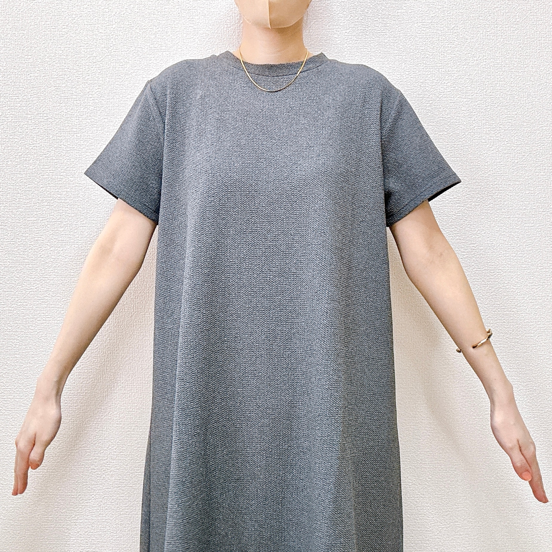 女性がシンプルなTシャツドレスを着て、腕を広げて立っている。シンプルなアクセサリーを身に着けている。