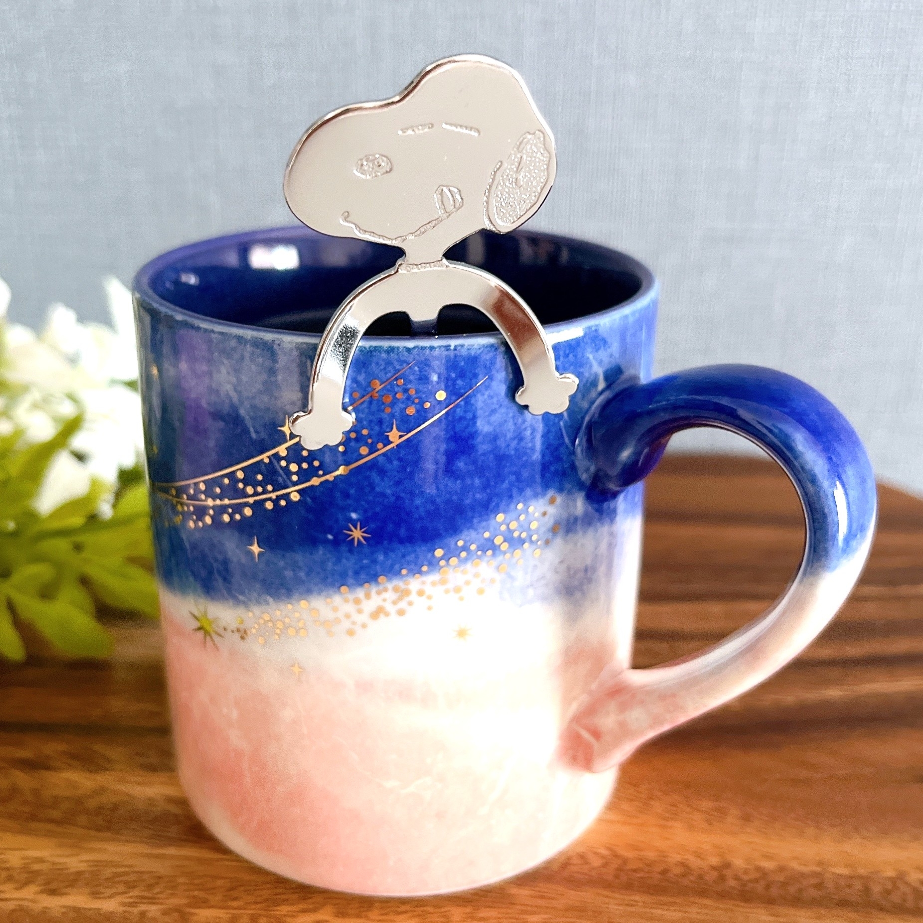 白い花の隣にあるカラフルなマグカップに、かわいい金属製のスヌーピーのティーバッグホルダーが引っかかっている。