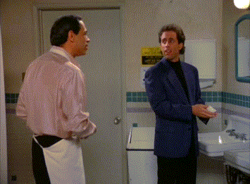 Two men in a restroom: one in a suit, the other in an apron. The suited man is holding a bar of soap, looking surprised. Scene appears comedic
