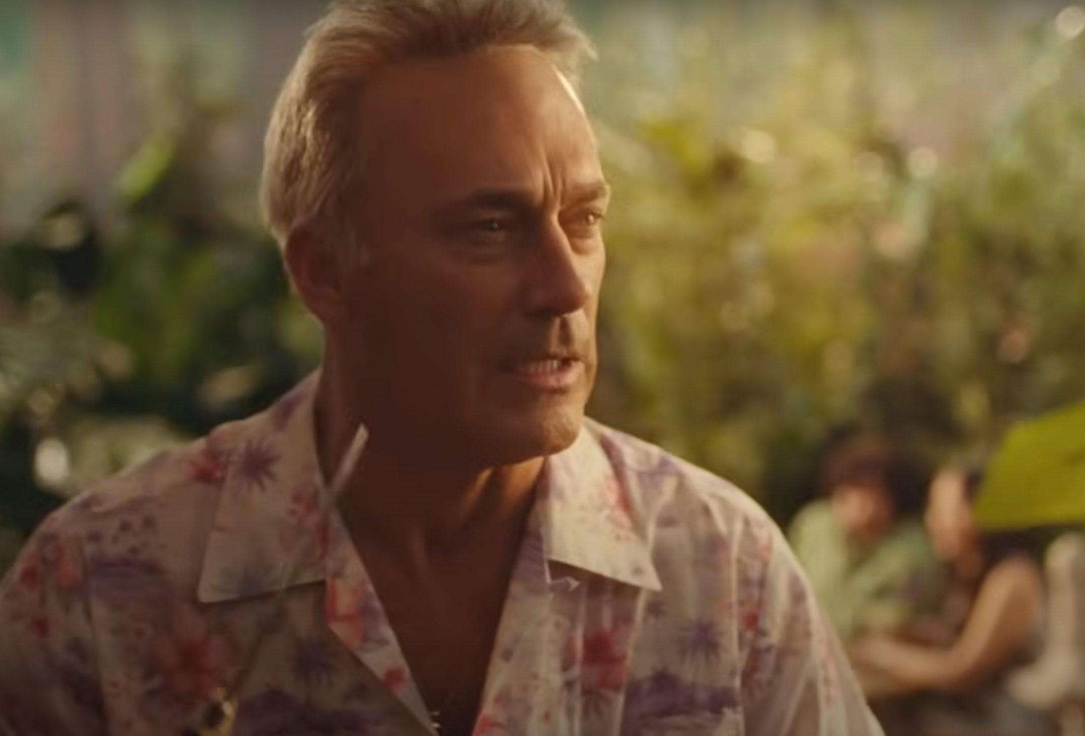 Man in a floral shirt looking to the side, with blurred greenery and people in the background