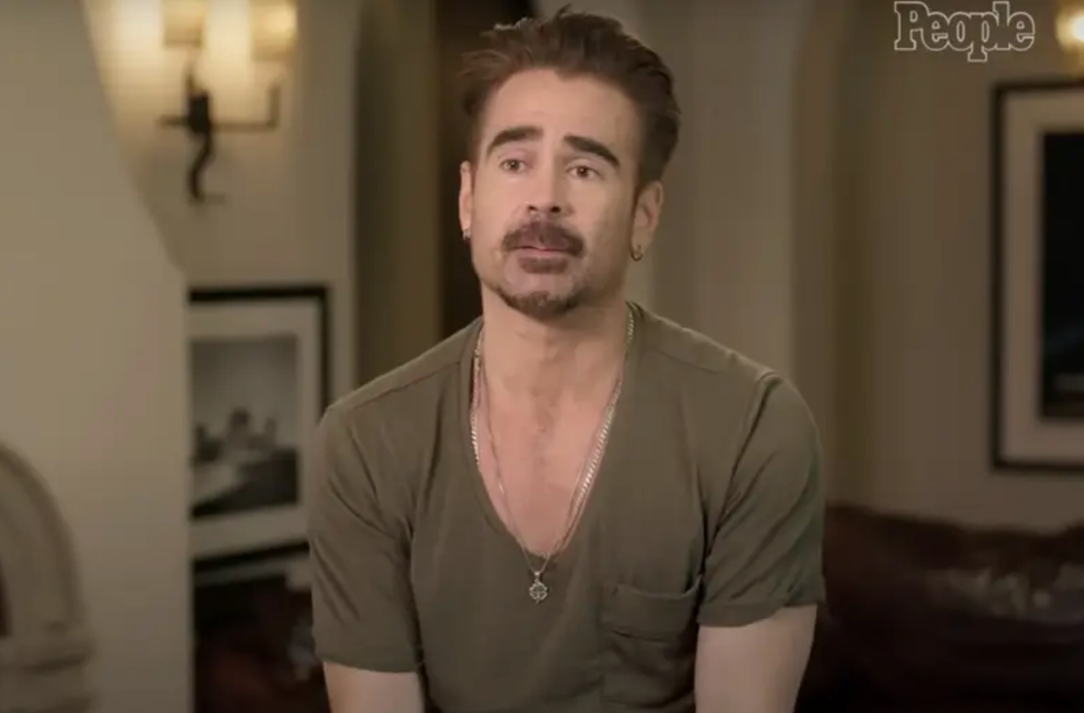 A person with facial hair wears a casual t-shirt, seated in an indoor setting with framed pictures on the wall