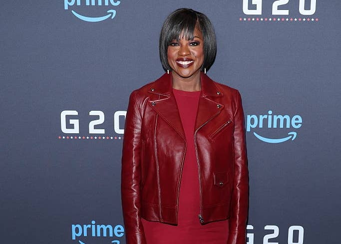 A person on the red carpet wearing a stylish red dress with a matching leather jacket and heels, standing in front of a Prime event backdrop