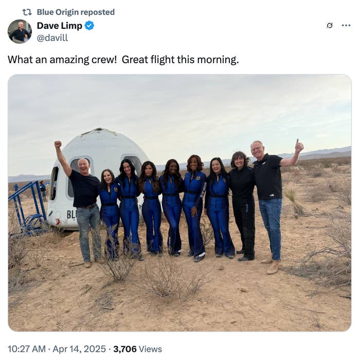 Group of people, including women in blue jumpsuits and others in casual wear, posing happily in front of a spacecraft on a desert-like terrain