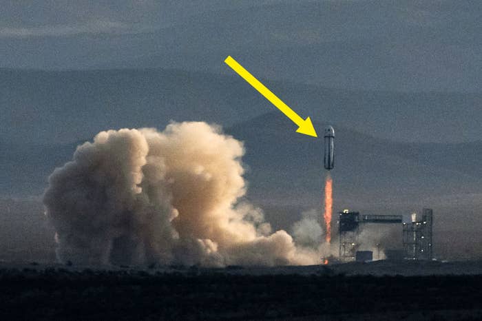 A rocket lifts off from a launchpad, emitting clouds of smoke and fire. Mountains are visible in the background