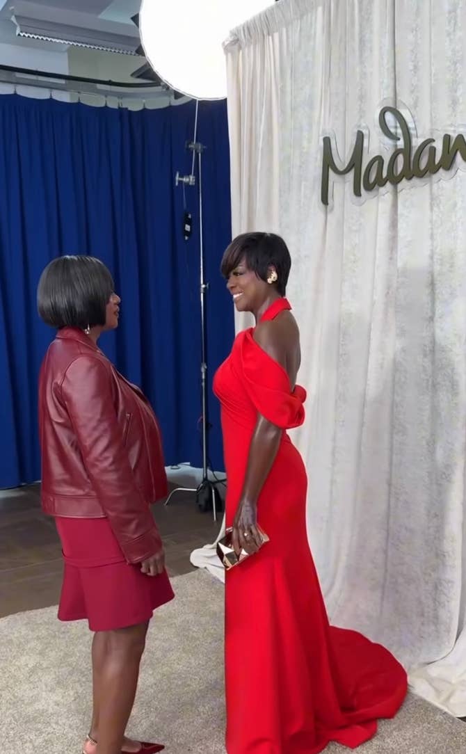 Two people conversing at an event. One wears a red gown with a halter neck design, holding a purse. The other wears a red jacket and skirt