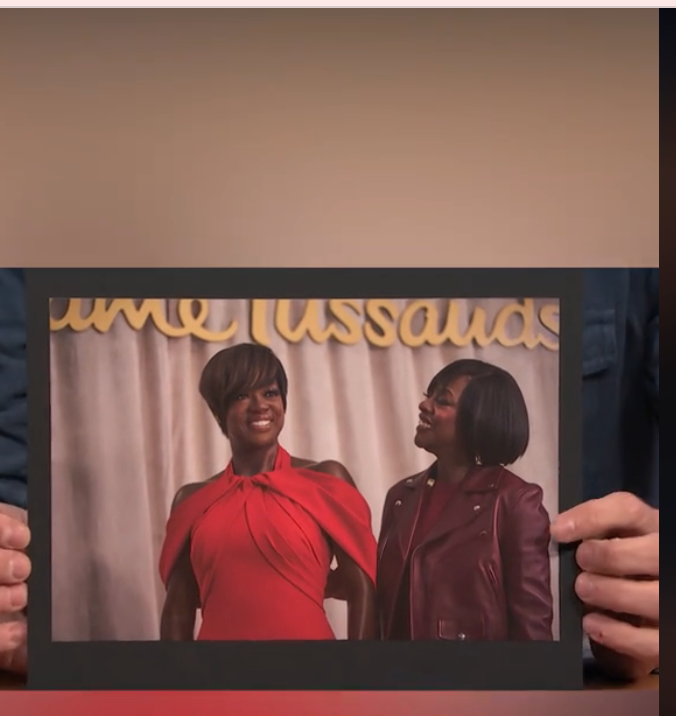 Person holds photo of a wax figure and a smiling individual. Figure wears an elegant, sleeveless dress, similar to person's stylish jacket