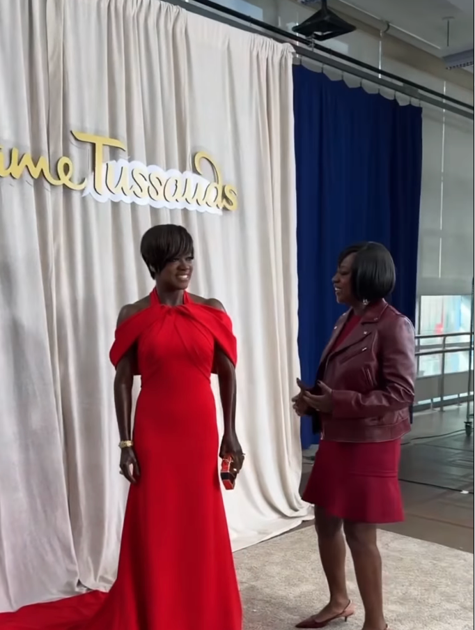 Two individuals stand in front of a Madame Tussauds sign. One wears a red gown with an off-the-shoulder style; the other wears a red jacket and skirt