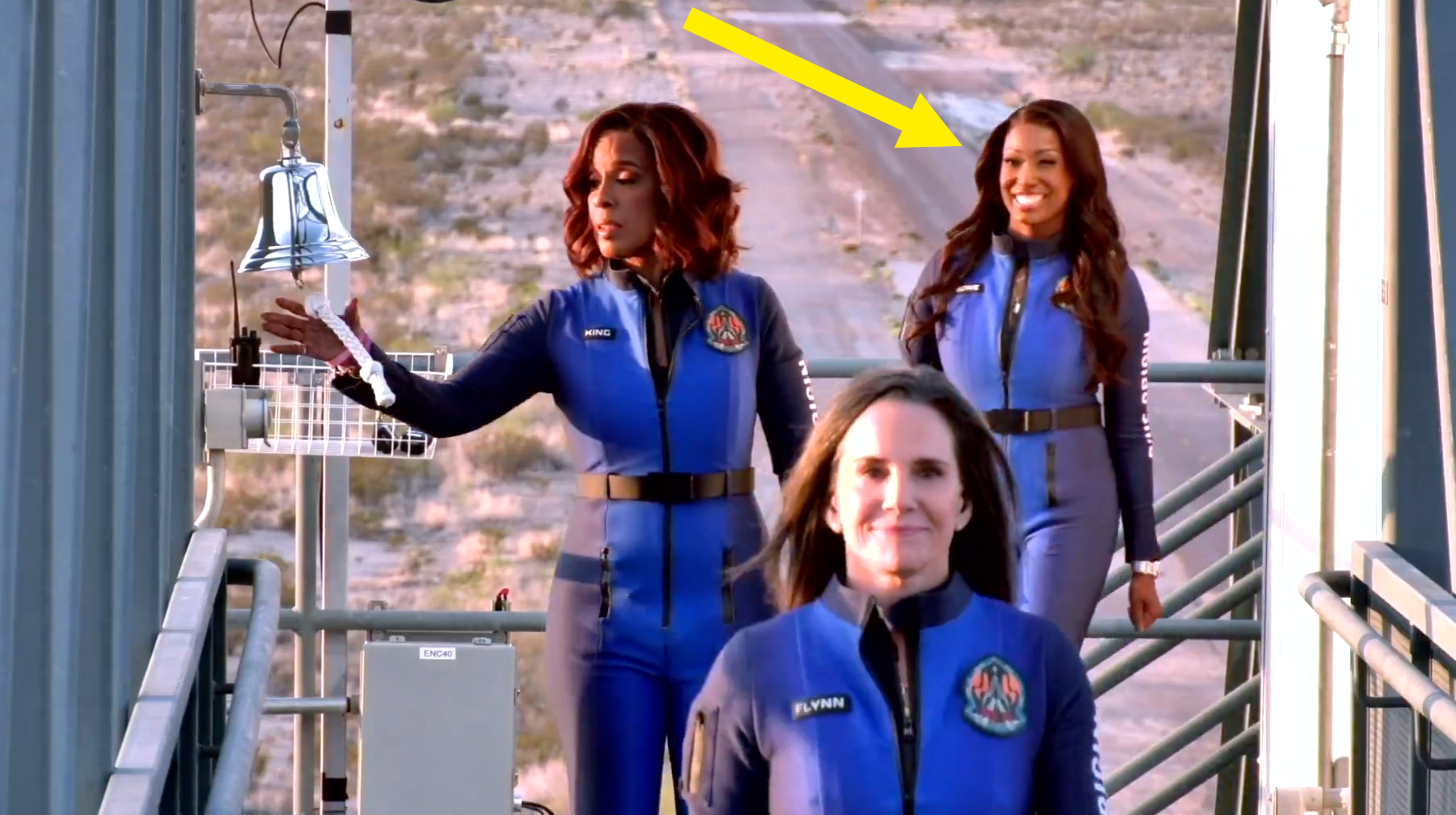 Three people in blue spaceflight suits walk on a launch pad, smiling, one about to ring a bell. The setting suggests a space exploration event