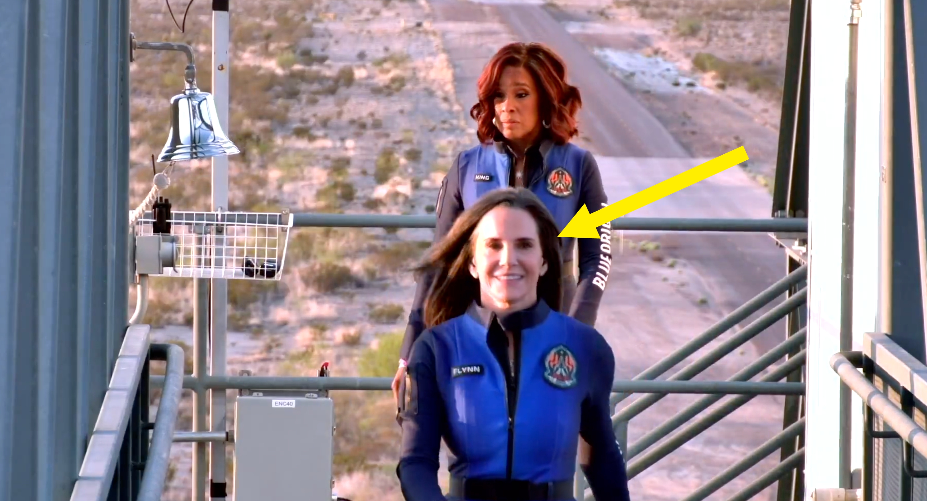 Two people dressed in blue astronaut jumpsuits walk on a platform, preparing for a space flight