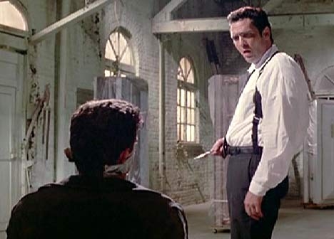 Two men in a tense standoff in a dim, industrial setting; one is standing with a knife. Scene from a movie