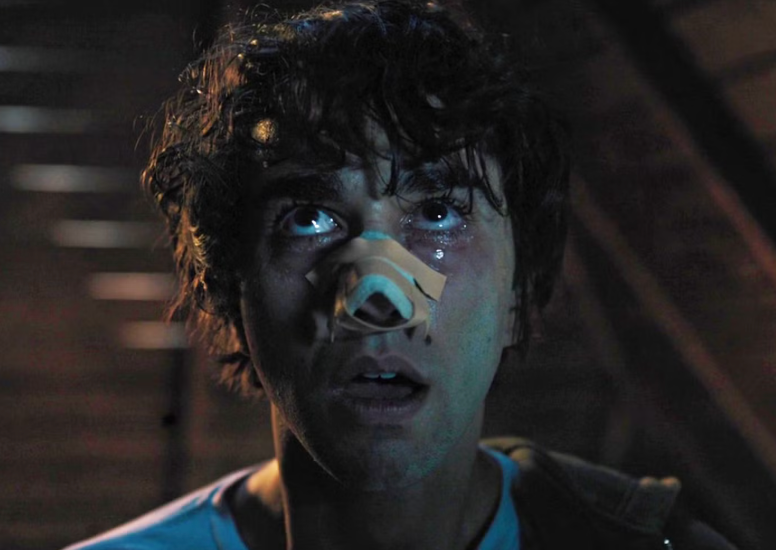 A person with a shocked expression, wearing a bandage on their nose, looks upwards in a dimly lit setting