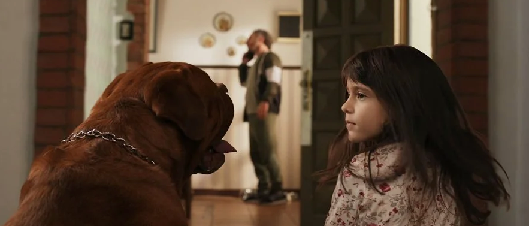 A young girl stands with a large dog in a hallway, watching a man in the background who is talking on the phone