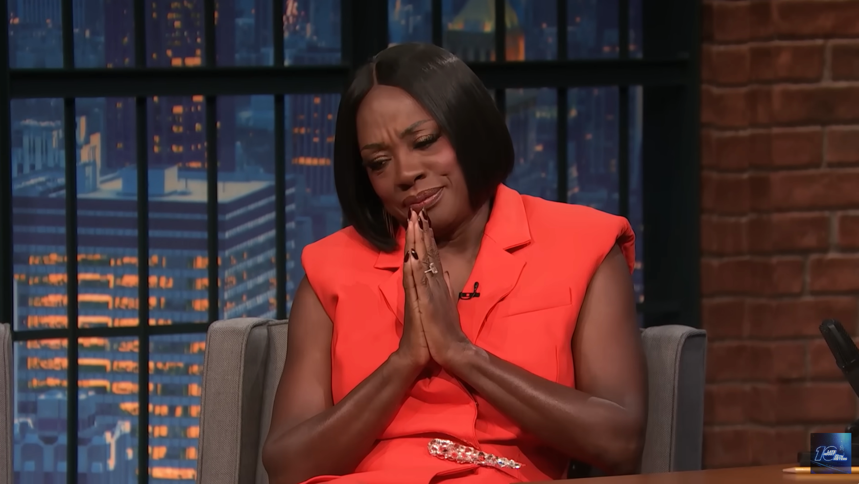 A celebrity on a talk show set, wearing a sleeveless, tailored dress, smiling and clasping hands, as if expressing gratitude or joy