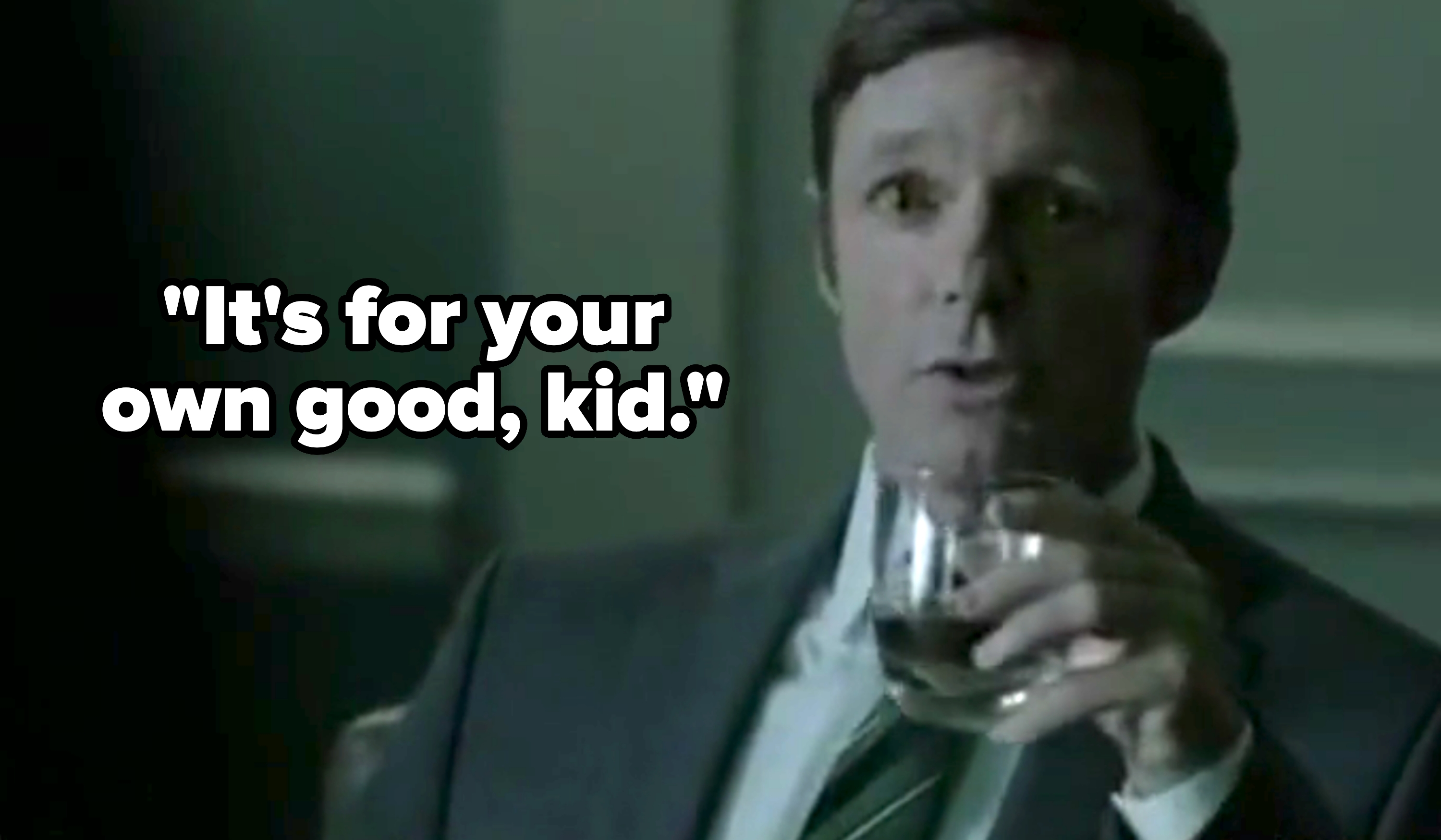 A person in a suit sits indoors holding a glass, appearing to be in conversation