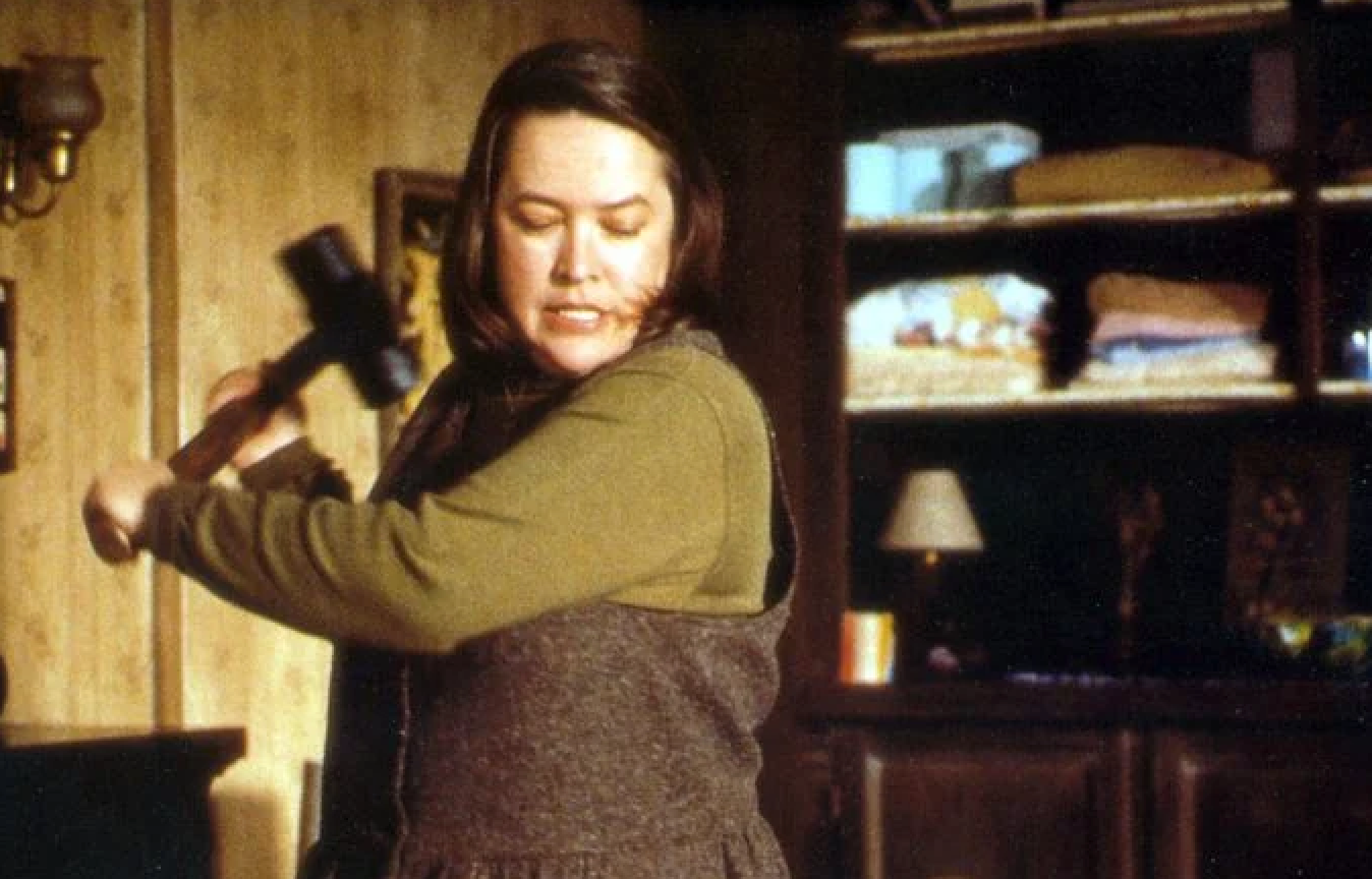 A person in a dramatic scene swinging a sledgehammer in an interior setting; shelves and a lamp in the background suggest a tense atmosphere