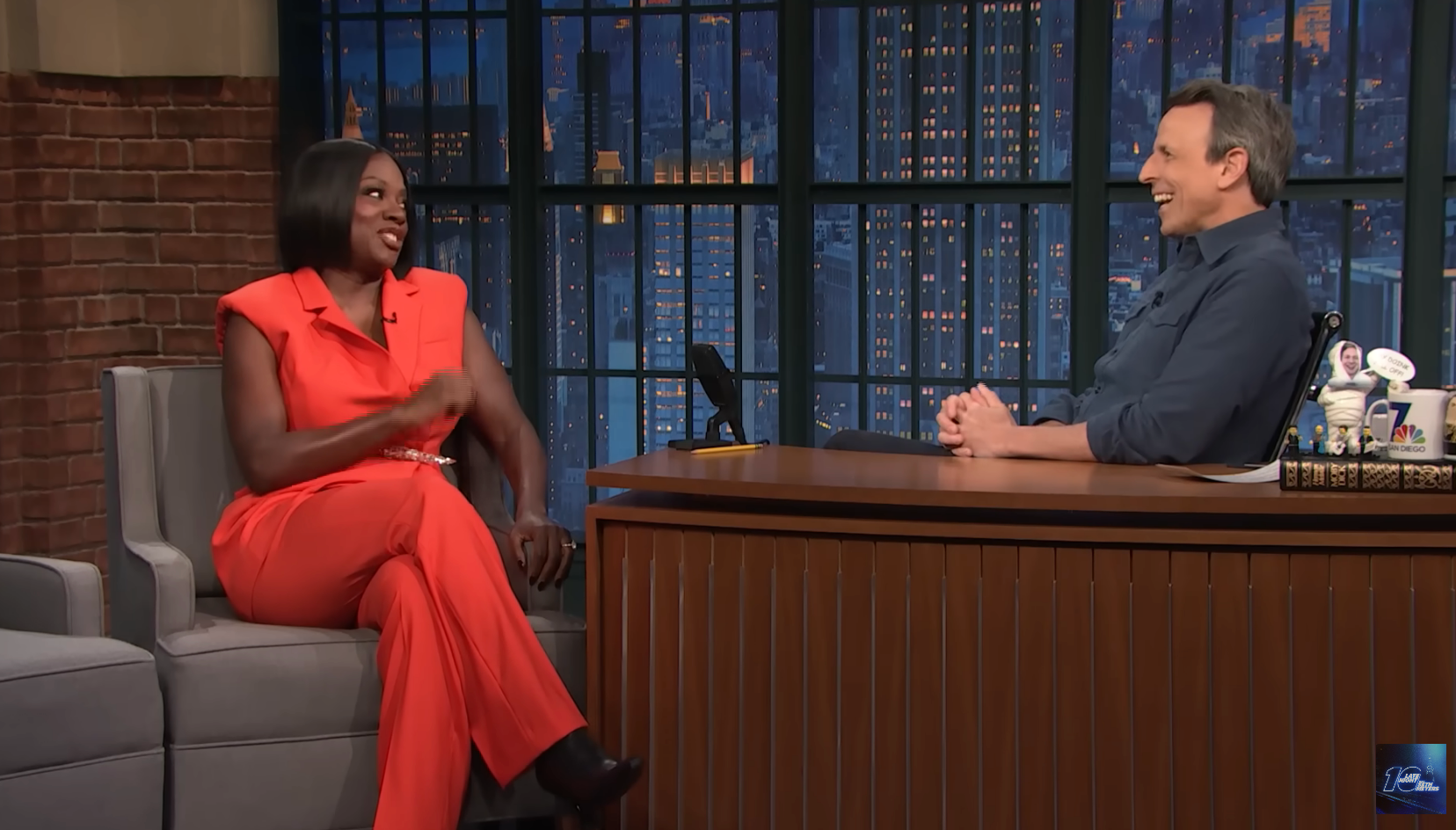 Talk show guest in elegant jumpsuit, animatedly conversing with host seated at a desk. Cozy studio setting with cityscape backdrop