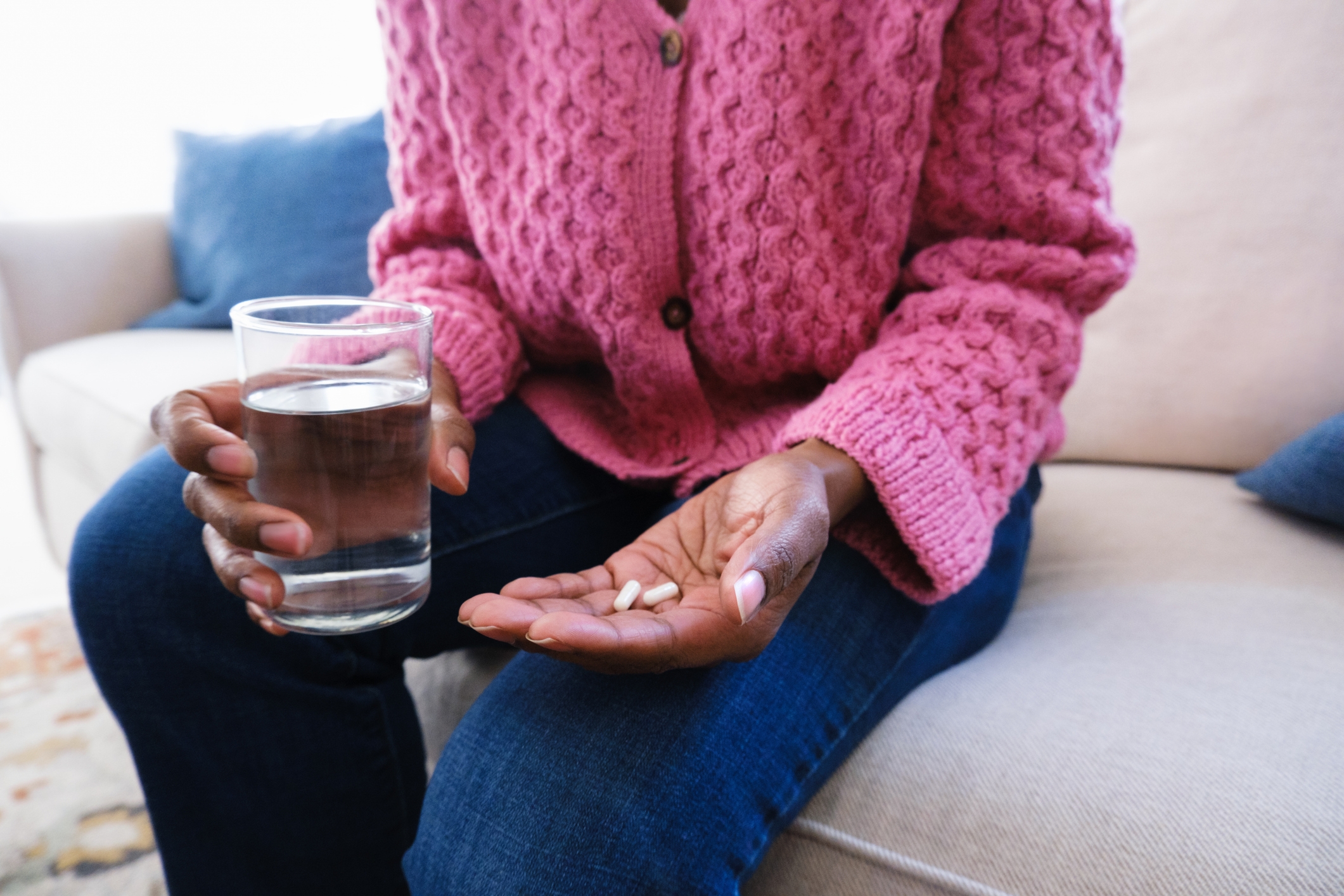 Person seated on a couch holding a glass of water and two pills in one hand, wearing a textured knit cardigan