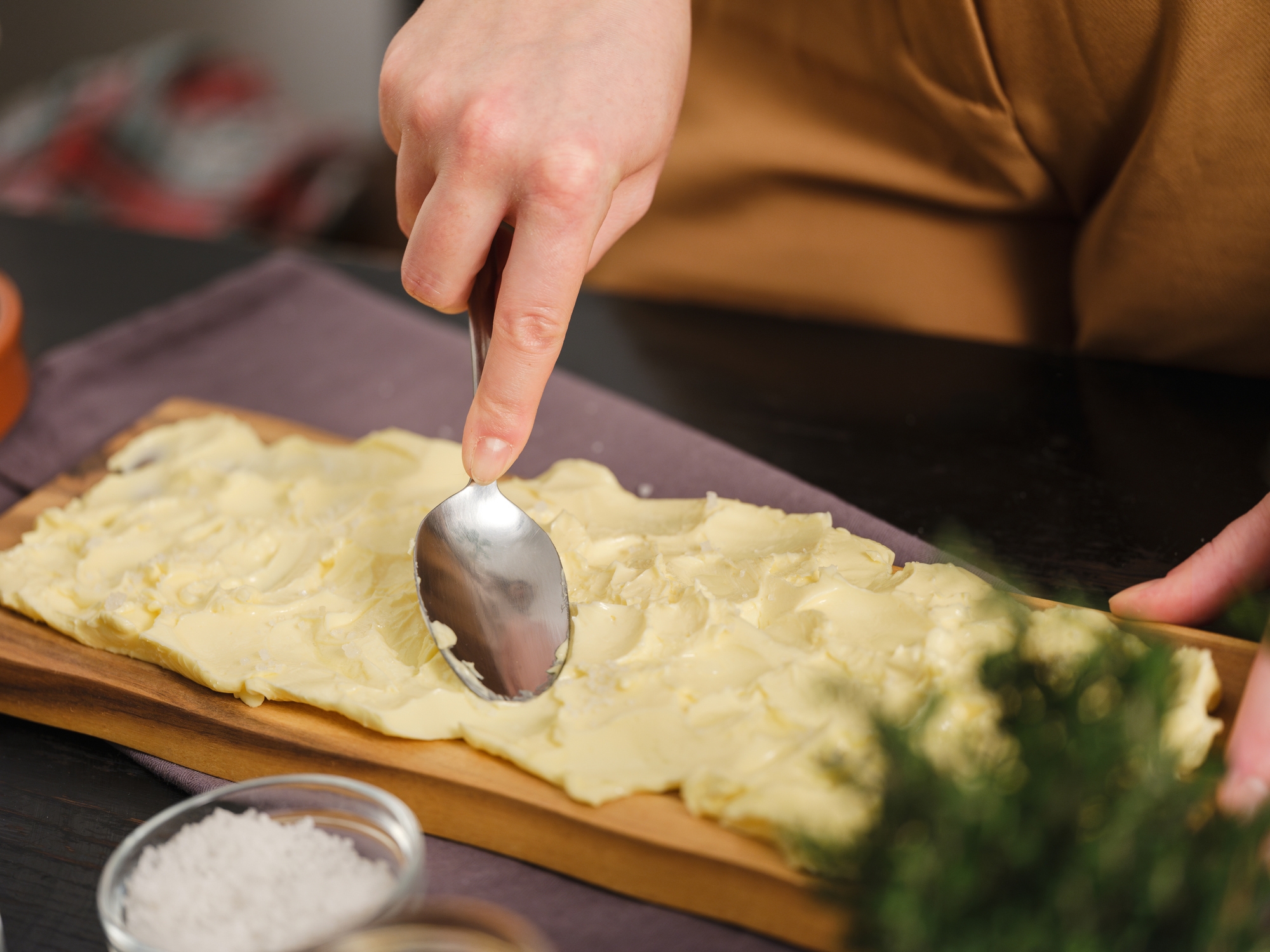 Person spreading butter on a wooden board using a spoon, with salt and herbs nearby