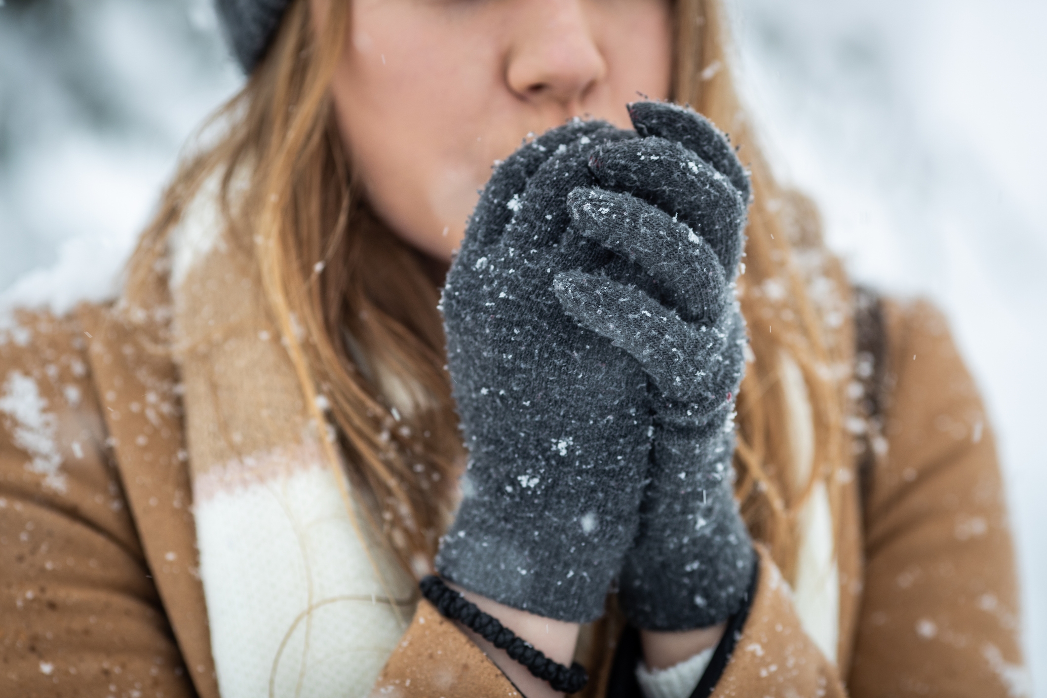 Person wearing gloves and a scarf blows into their hands for warmth in a snowy setting