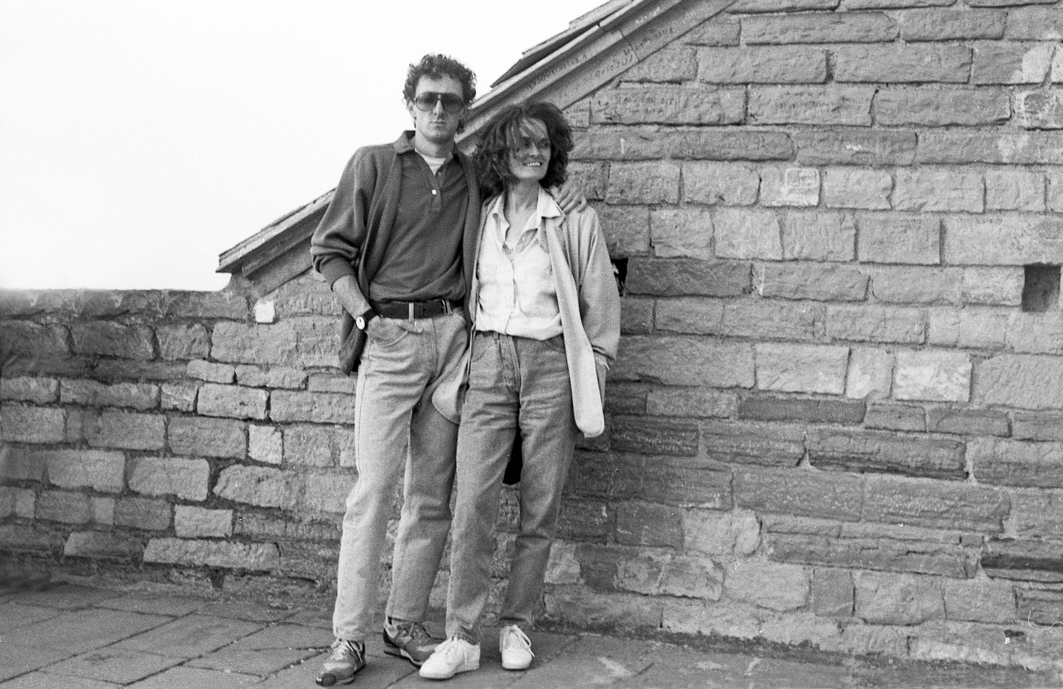 Two people in casual clothes stand close together against a stone wall, both with hands in their pockets and smiling at the camera